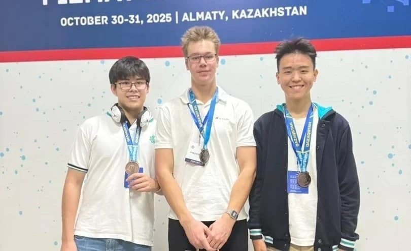 Three students wearing FAIO 2025 bronze medals in front of the olympiad banner in Almaty